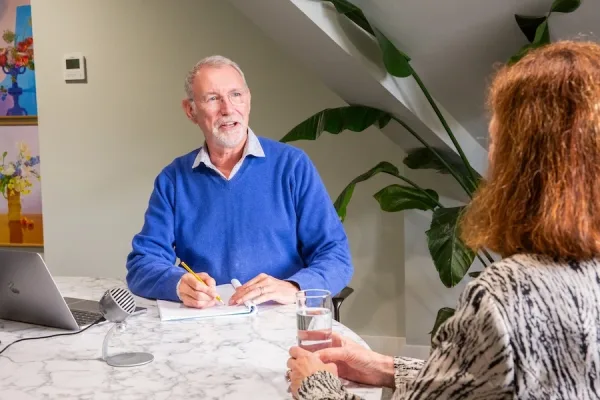 Andrew Gent sitting at a table with a client, writing down what she's saying. 